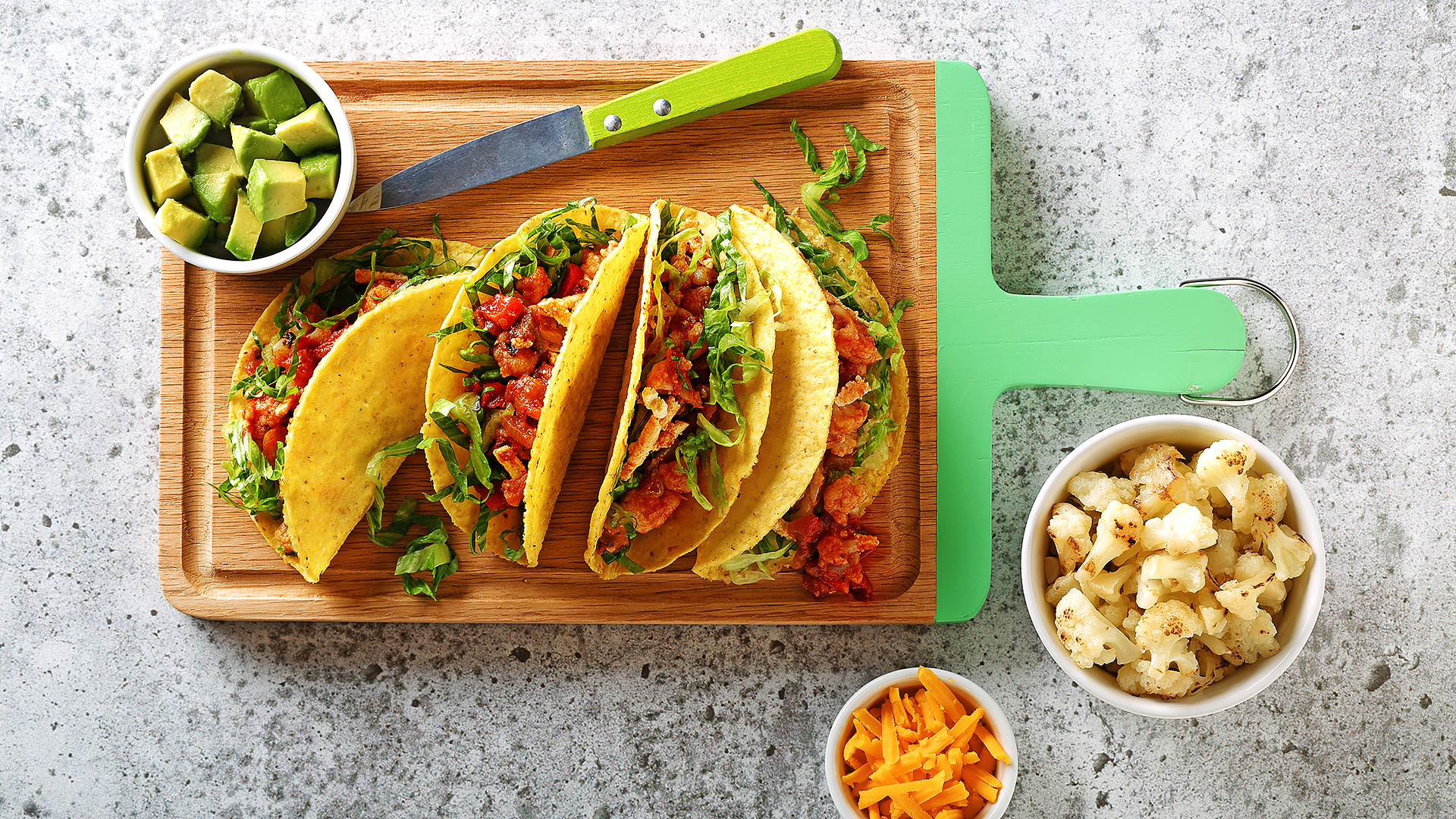 Cauliflower tacos displayed on a cutting board with side bowls filled with cheese, avocado and cauliflower. 