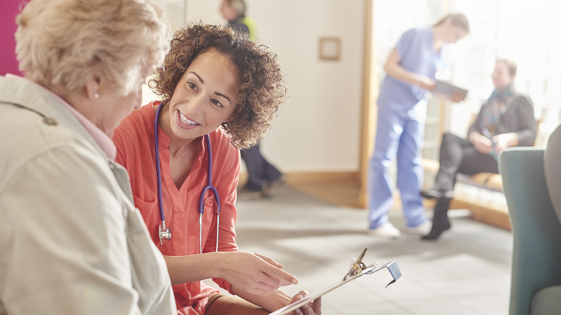 A female doctor stands next to a senior patient in her waiting room and casually chats to her about her details. In the background a nurse with a tablet and a patient at the desk and booking an appointment.